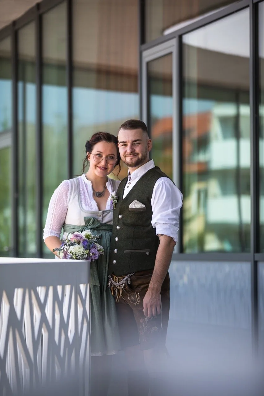 Brautpaar in Tracht vor einer Glasfassade, davor der Brautstrauß