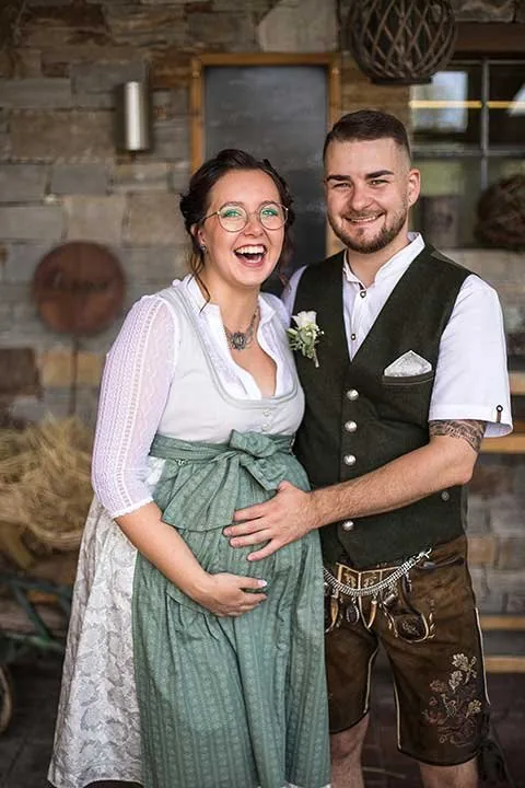 Brautpaar in Tracht lacht Arm in Arm vor einer Steinwand