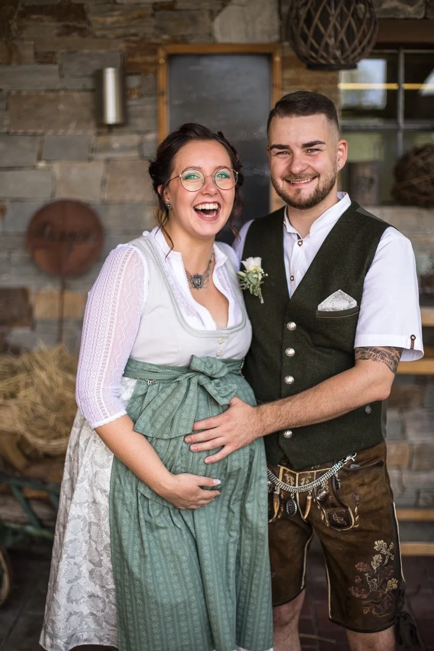 Brautpaar in Tracht lacht Arm in Arm vor einer Steinwand