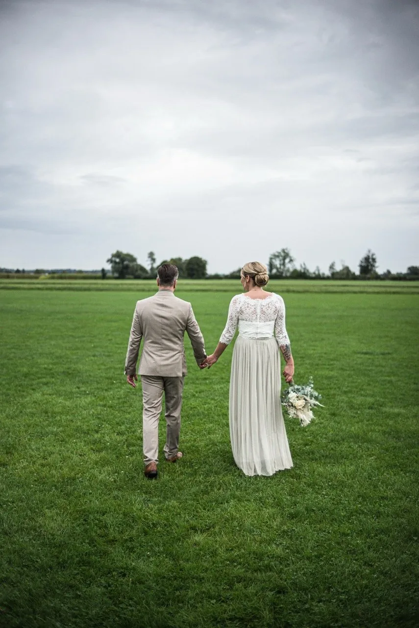 Brautpaar läuft Hand in Hand über eine weite Wiese, von hinten fotografiert