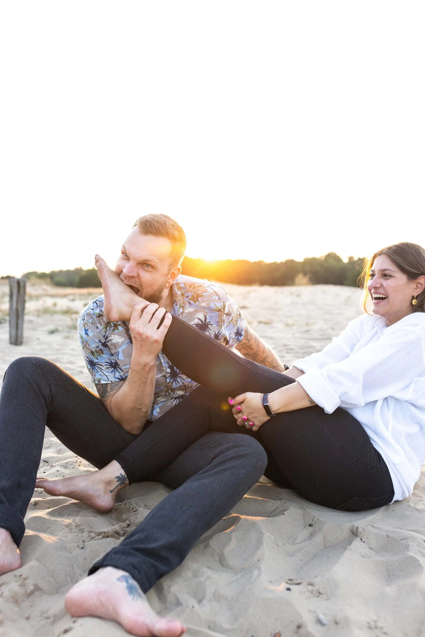 Das Paar sitzt lachend im Sand, die Sonne steht tief über dem Waldrand